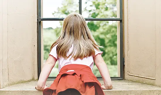 Ein Kind mit blonden langen Haaren ist von hinten zu sehen, während es durch ein großes Fenster mit Metallrahmen ins Grüne guckt.