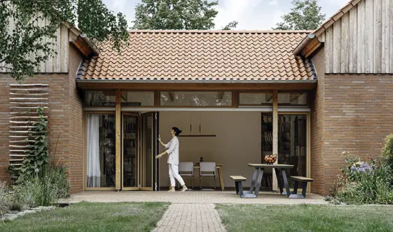 Frau öffnet Glas-Faltwand mit Holzrahmen eines Ziegelsteinhauses zur gepflasterten Terrasse im Garten.